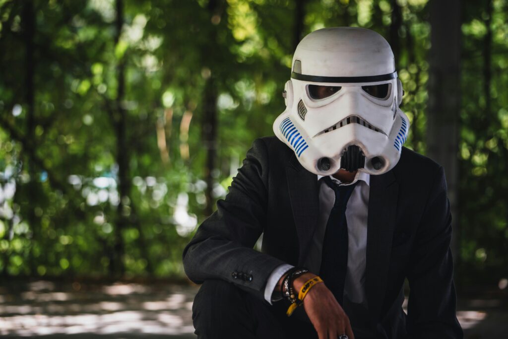A person in a suit wearing a stormtrooper helmet outdoors in a shaded area.